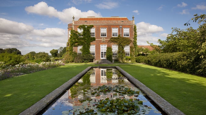 Georgian-style red brick house covered in climbing ivy, viewed from a formal garden with a long rectangular reflecting pool filled with lily pads in the foreground, mirrored in the still water, under a bright blue sky with scattered clouds.